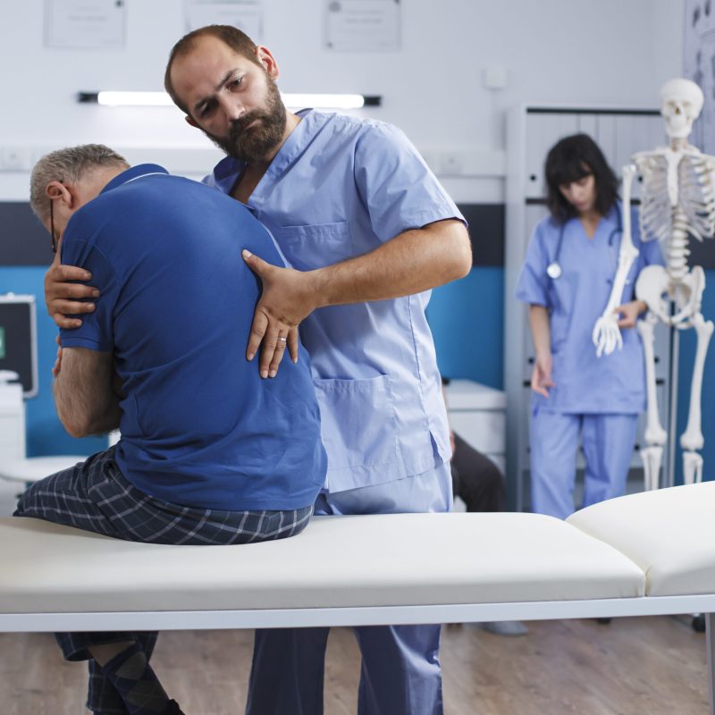 Osteopathic nurse gently extends spine of elderly patient with injury as part of physiotherapy. Chiropractor provides aid to gentleman experiencing back pains, aiding in physical recovery.