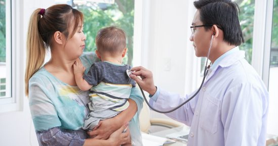 Asian young mother holding her toddler on her arms while male pediatrician listening to him with stethoscope